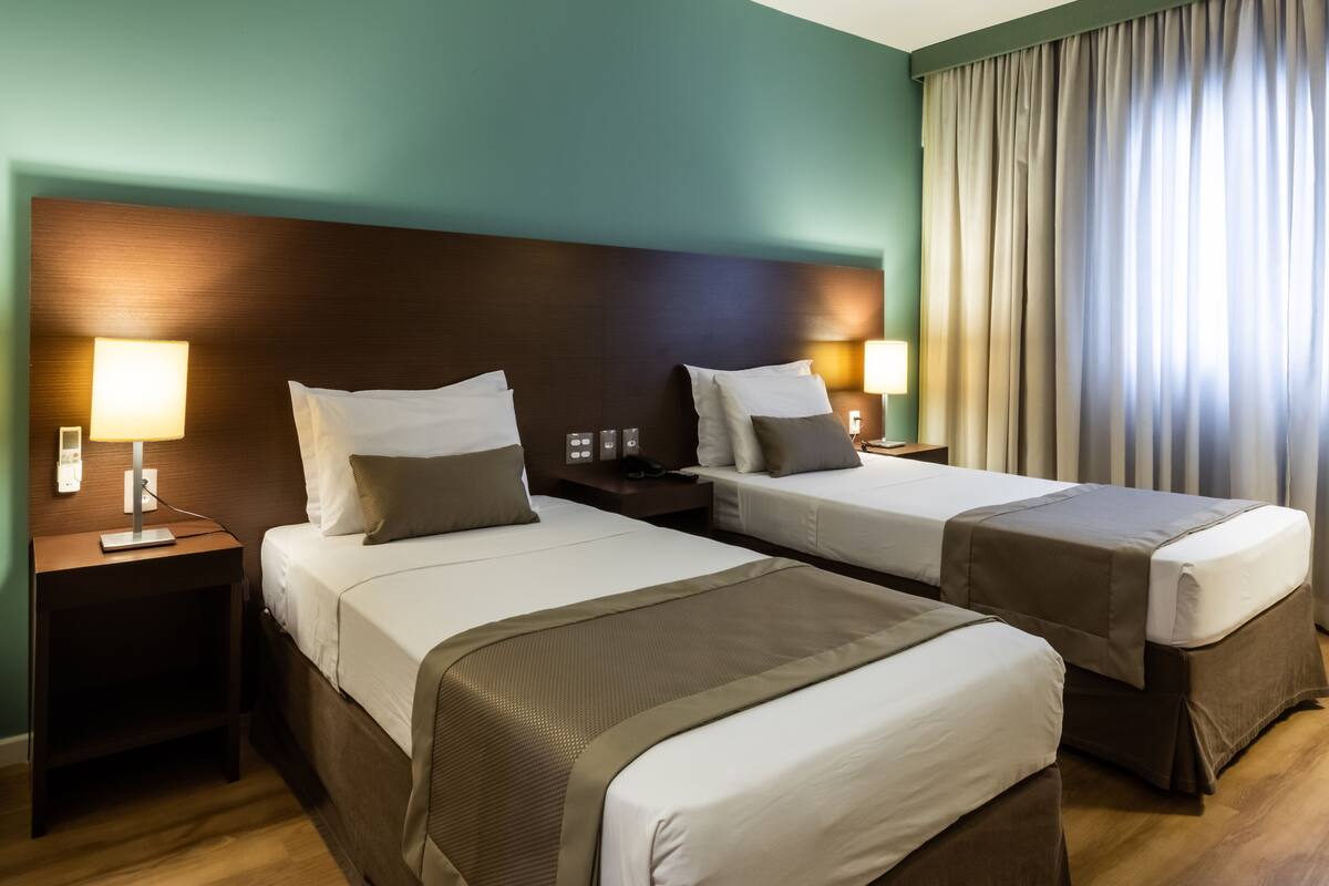 A hotel room with two beds, nightstands with lamps, a wooden headboard, and a large window with curtains.