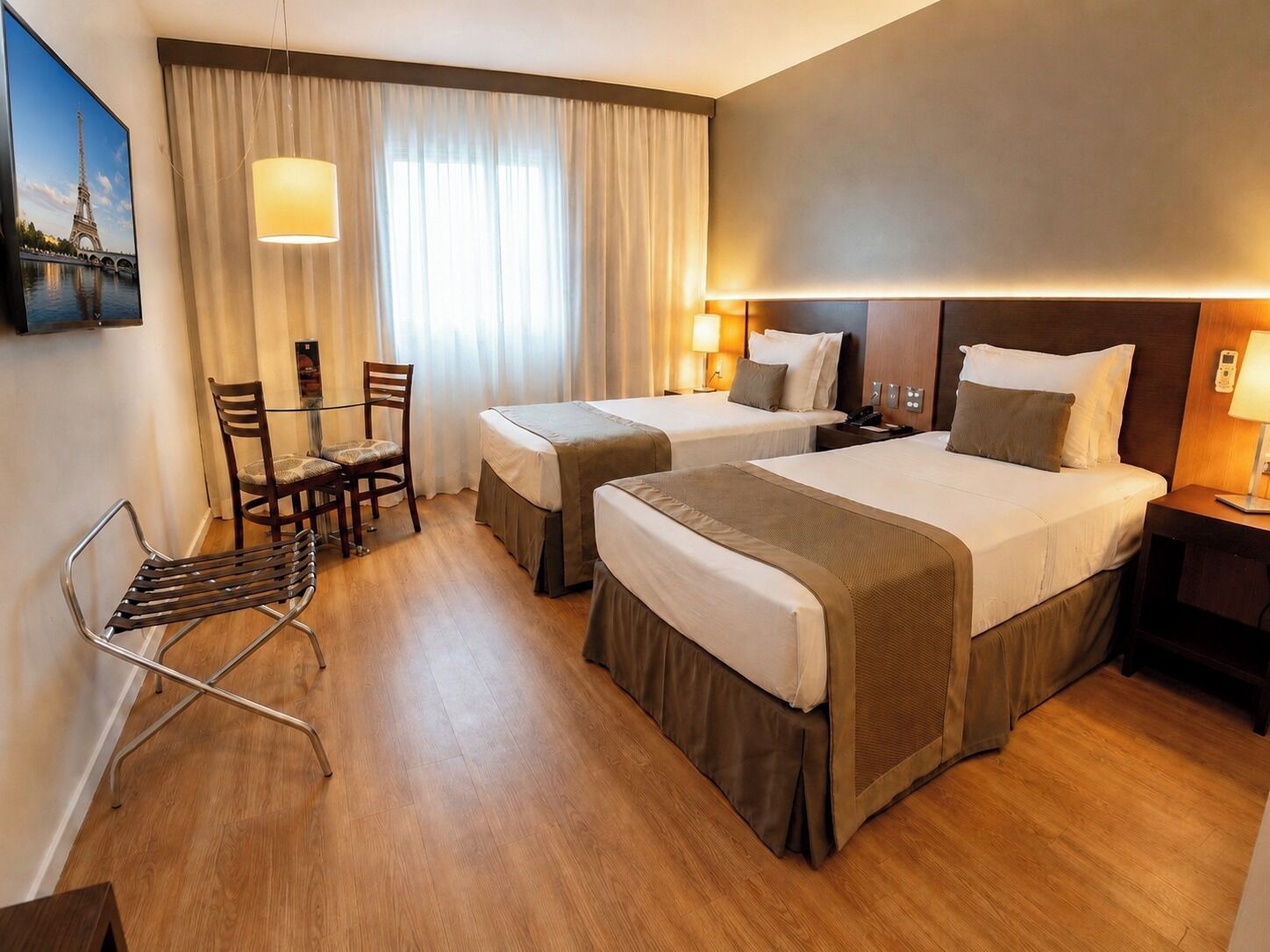 A hotel room with two beds, a desk, a chair, a lamp, and a painting of the Eiffel Tower.