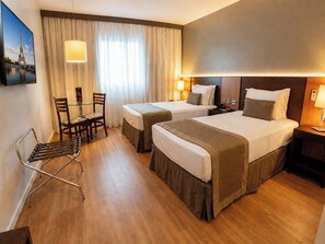 Une chambre d’hôtel avec deux lits, un bureau, une chaise, une lampe et un tableau représentant la Tour Eiffel.