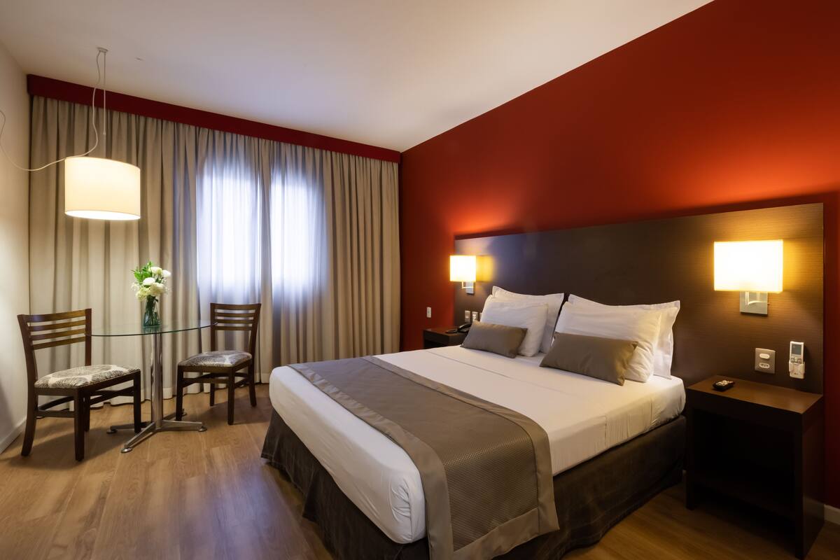 A hotel room with a large bed, two bedside tables, a glass table with chairs, and a view of the outside through curtains.