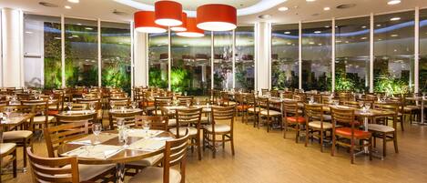 A spacious restaurant with multiple dining tables and chairs, large windows with greenery, and modern ceiling lights.