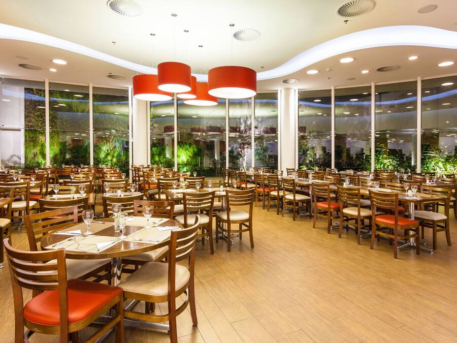 Ein geräumiges Restaurant mit mehreren Tischen und Stühlen, großen Fenstern mit Grünpflanzen und modernen Deckenleuchten.