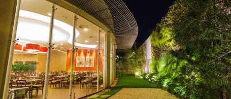 A modern building with a glass facade, a well-lit interior, and a pathway leading to a green area with trees.