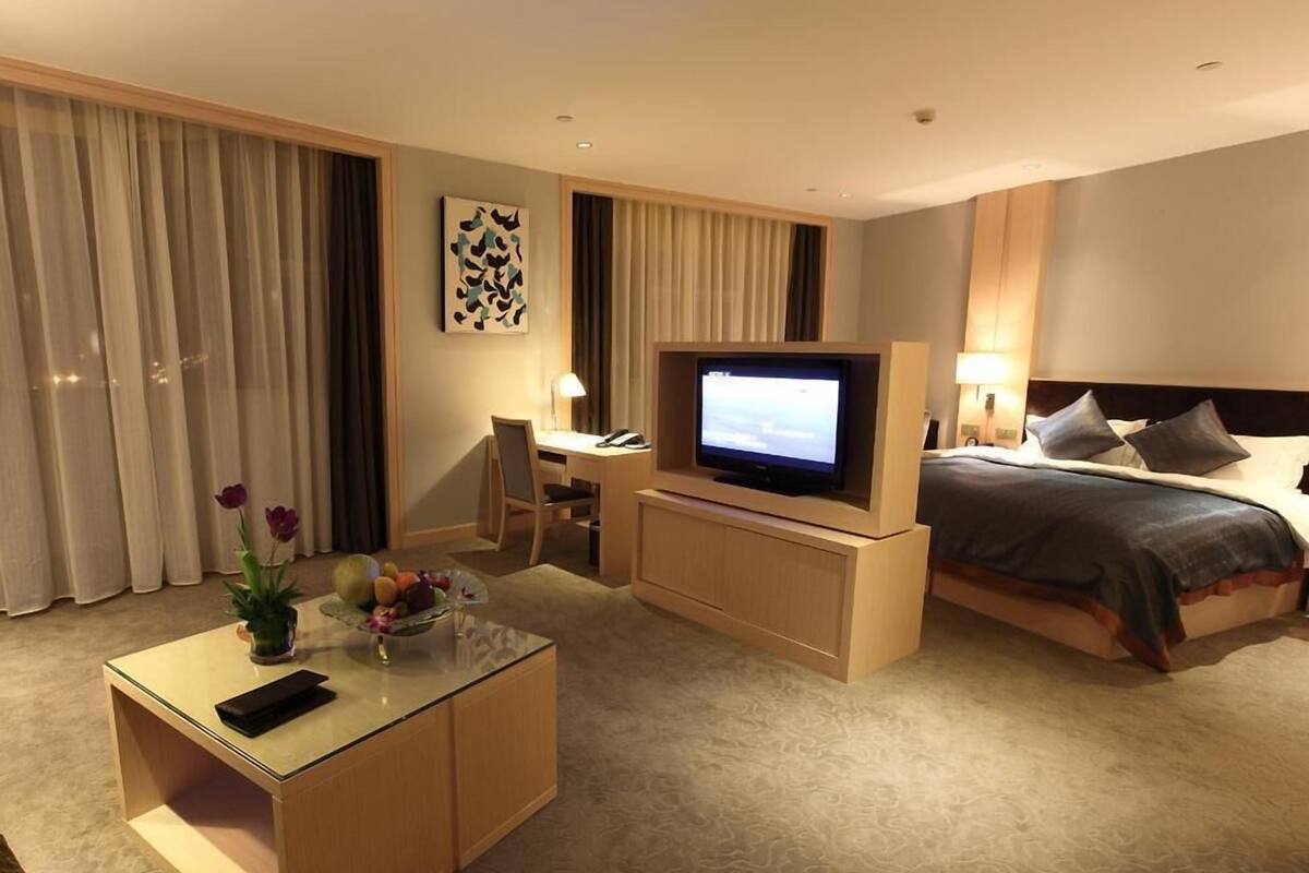 A hotel room with a bed, a desk with a TV, a small table with a vase, and a chair.