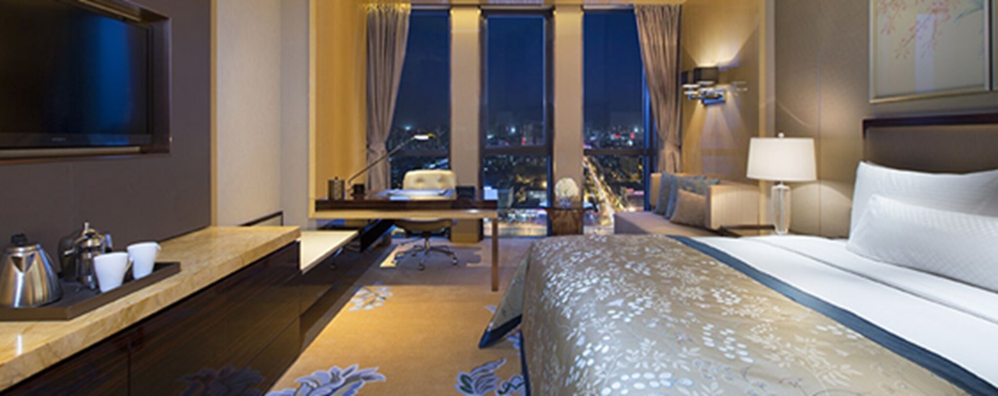 A hotel room with a bed, a desk, a chair, a lamp, and a view of the city at night.