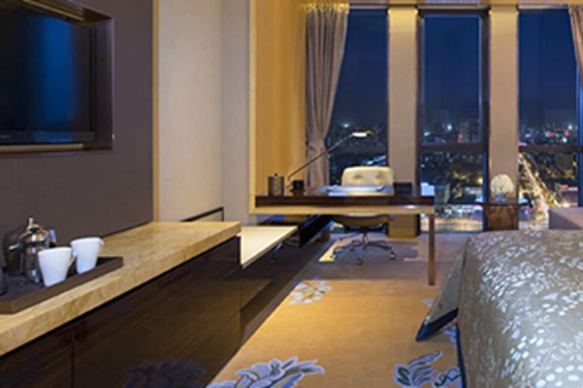 A hotel room with a bed, a desk, a chair, a lamp, and a view of the city at night.