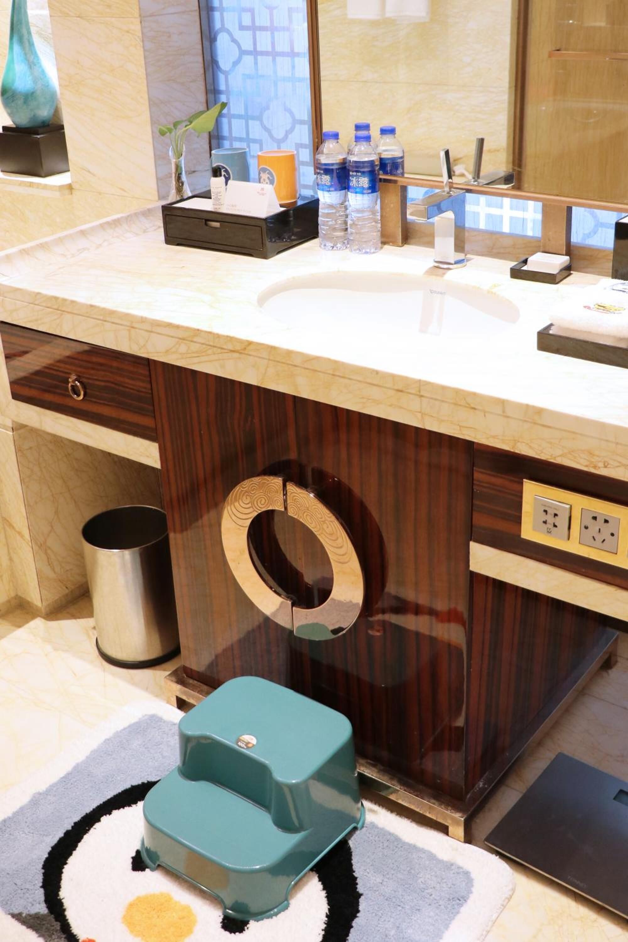 A bathroom with a marble sink, a wooden vanity, a trash can, a scale, and a green step stool.