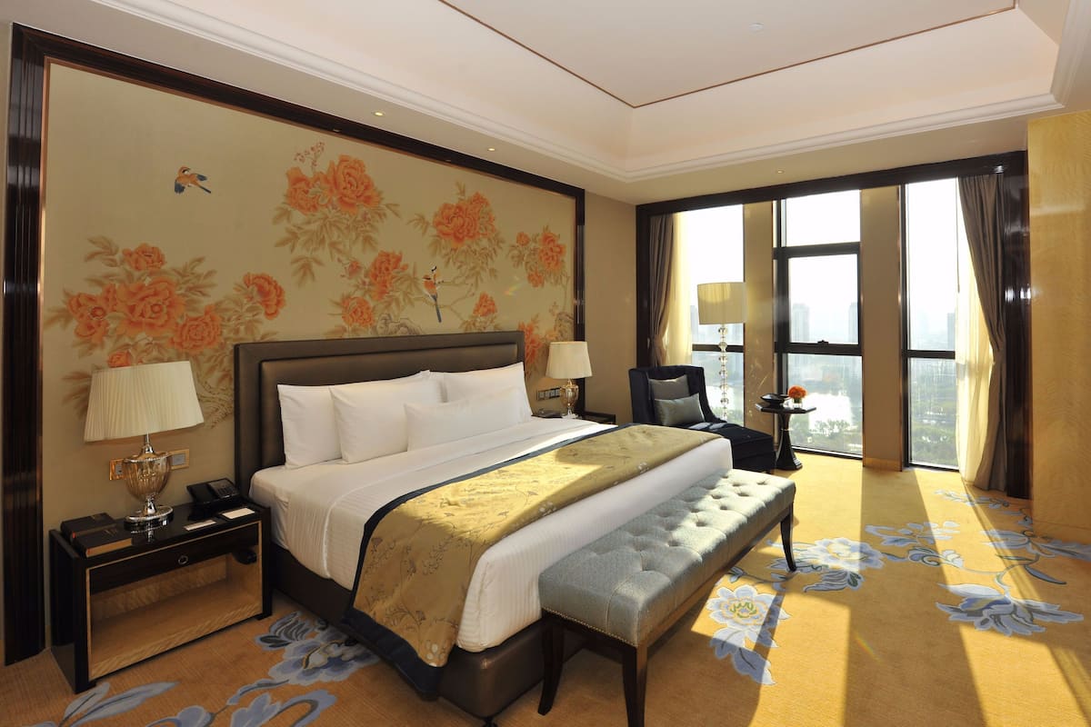 A hotel room with a large bed, a bench, a nightstand, a lamp, and a view of the city through large windows.
