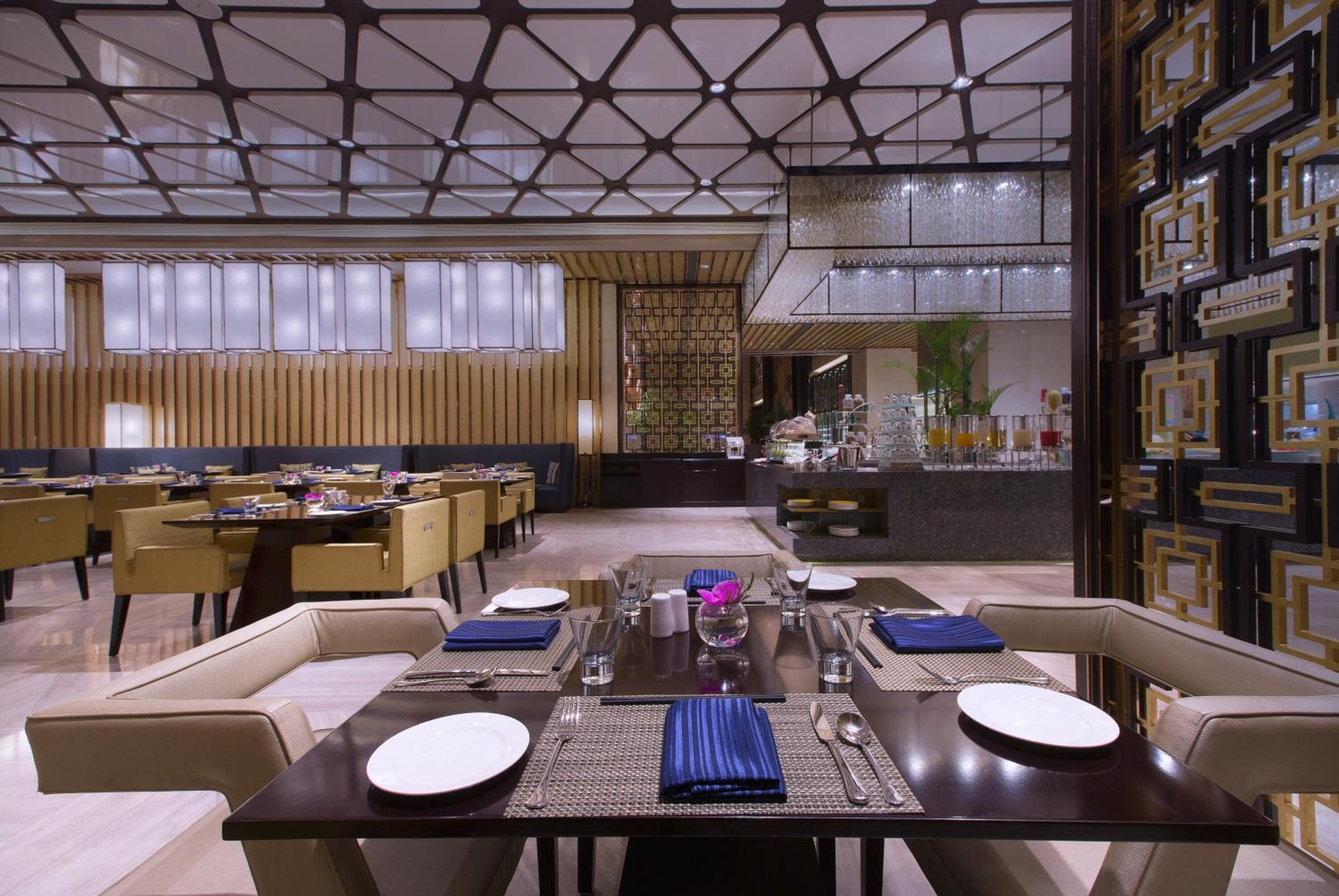 A modern dining area with a geometric ceiling design, multiple tables set for dining, and a bar area in the background.