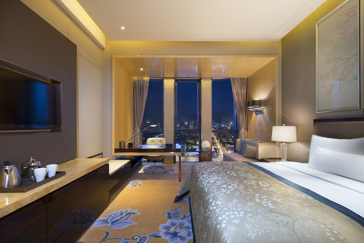 A hotel room with a bed, a desk with a chair, a TV, a nightstand with a lamp, and a view of the city at night.