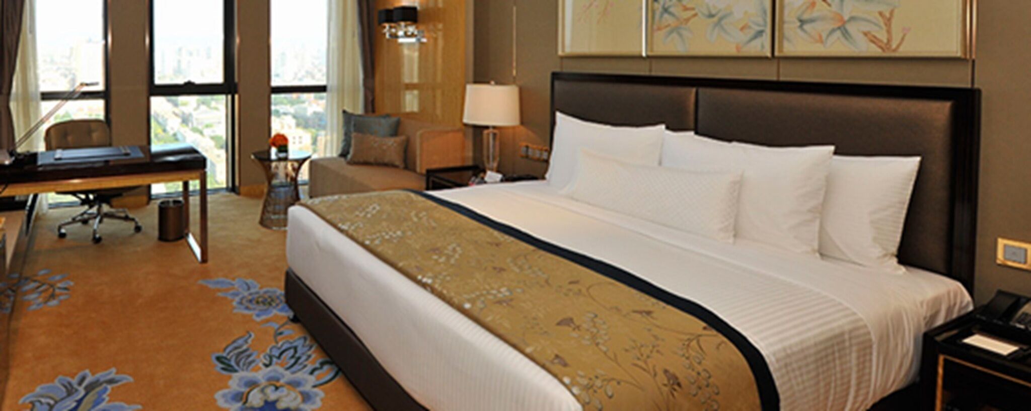 A hotel room with a large bed, a desk, a chair, a lamp, and a view of the city through the window.
