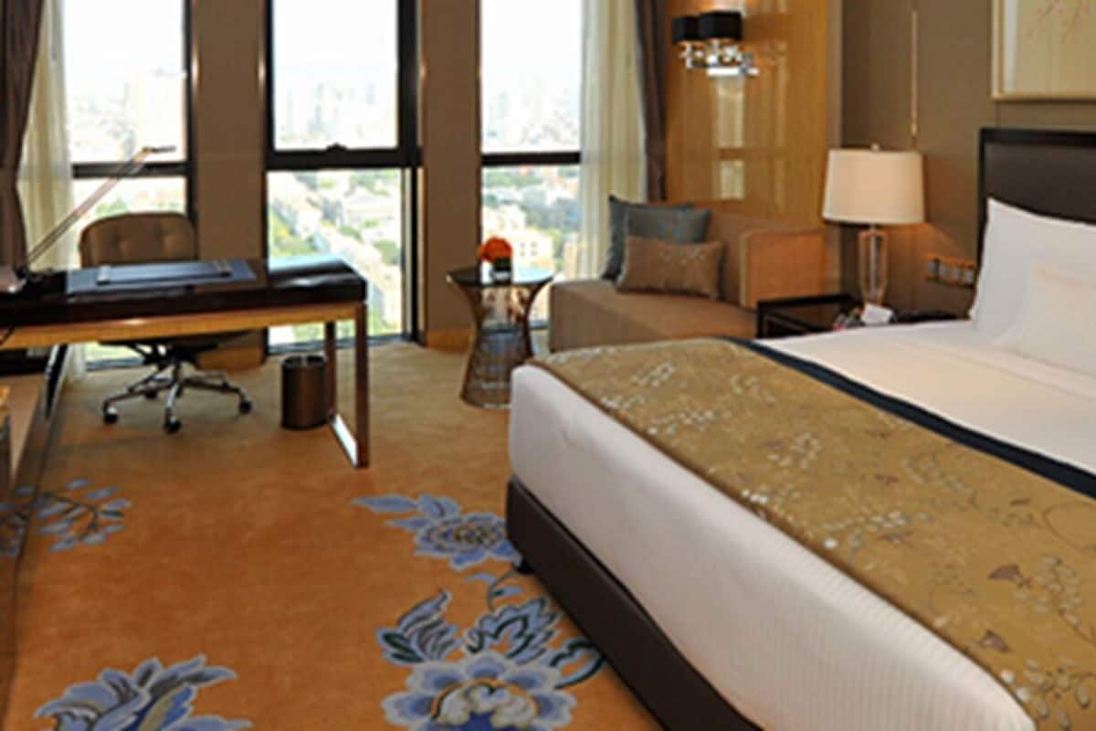A hotel room with a large bed, a desk, a chair, a lamp, and a view of the city through the window.