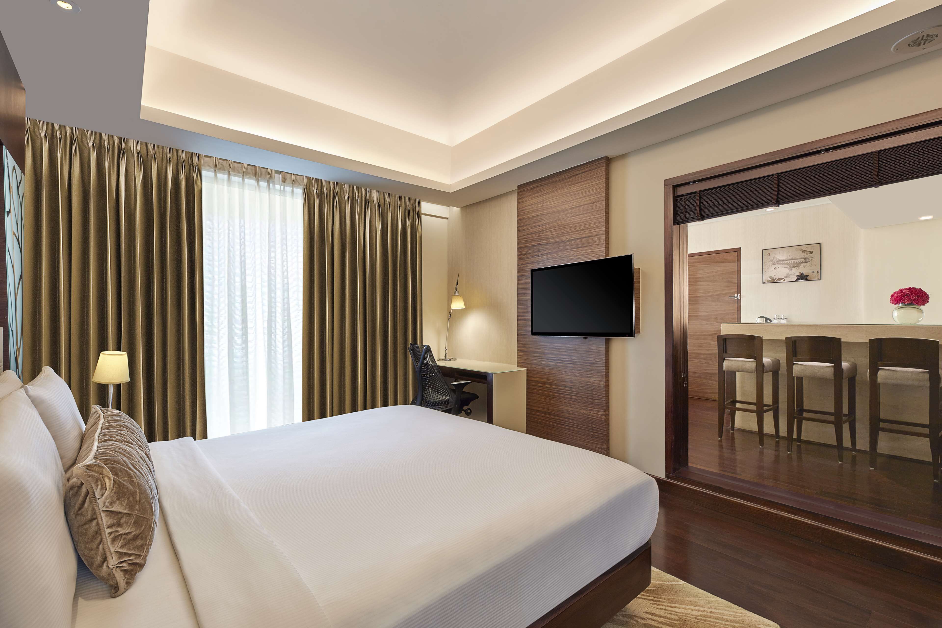 A modern hotel room with a large bed, a desk, a flat-screen TV, and a small bar area with stools.