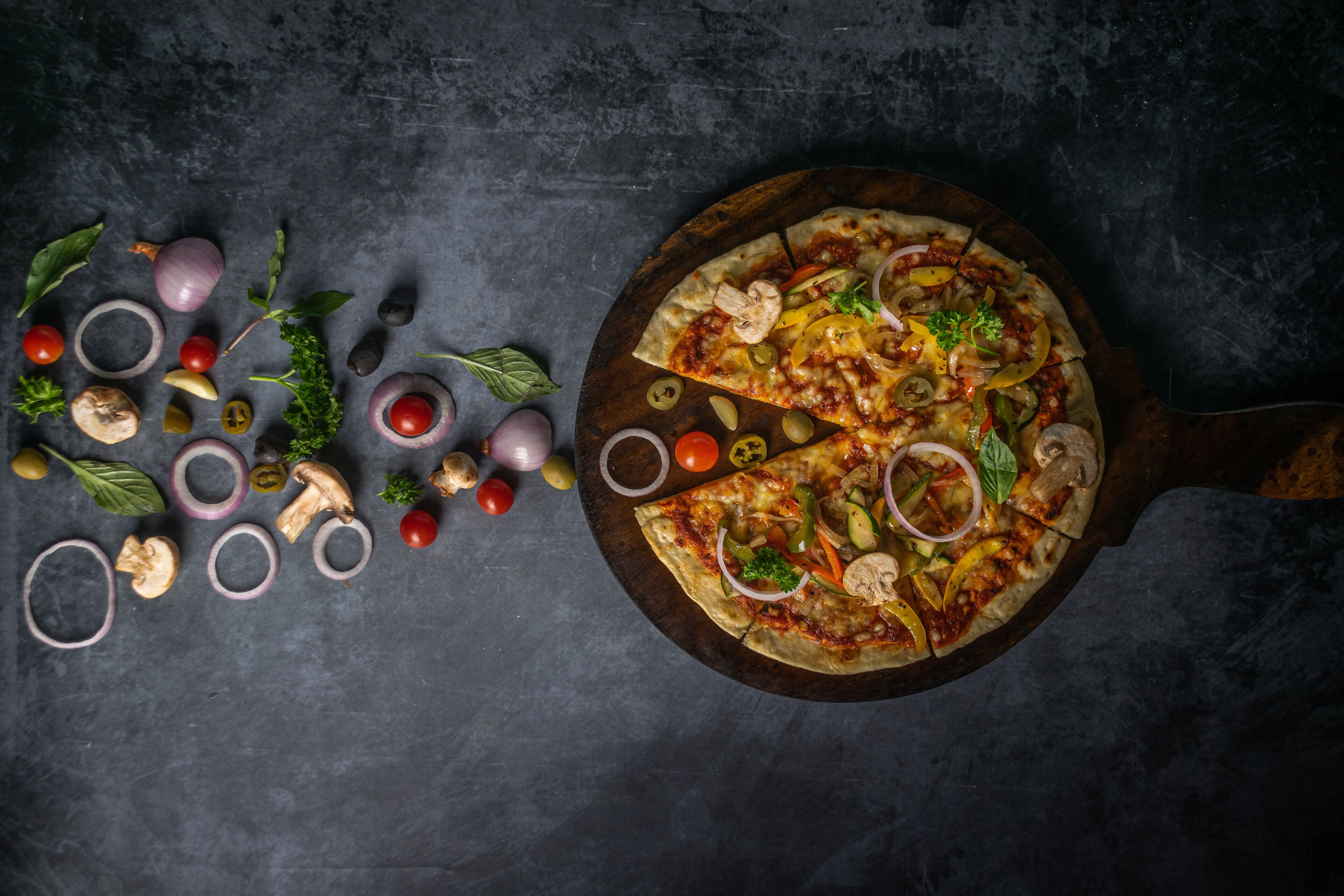 Una pizza sobre una tabla de madera, rodeada de diferentes verduras y hierbas.