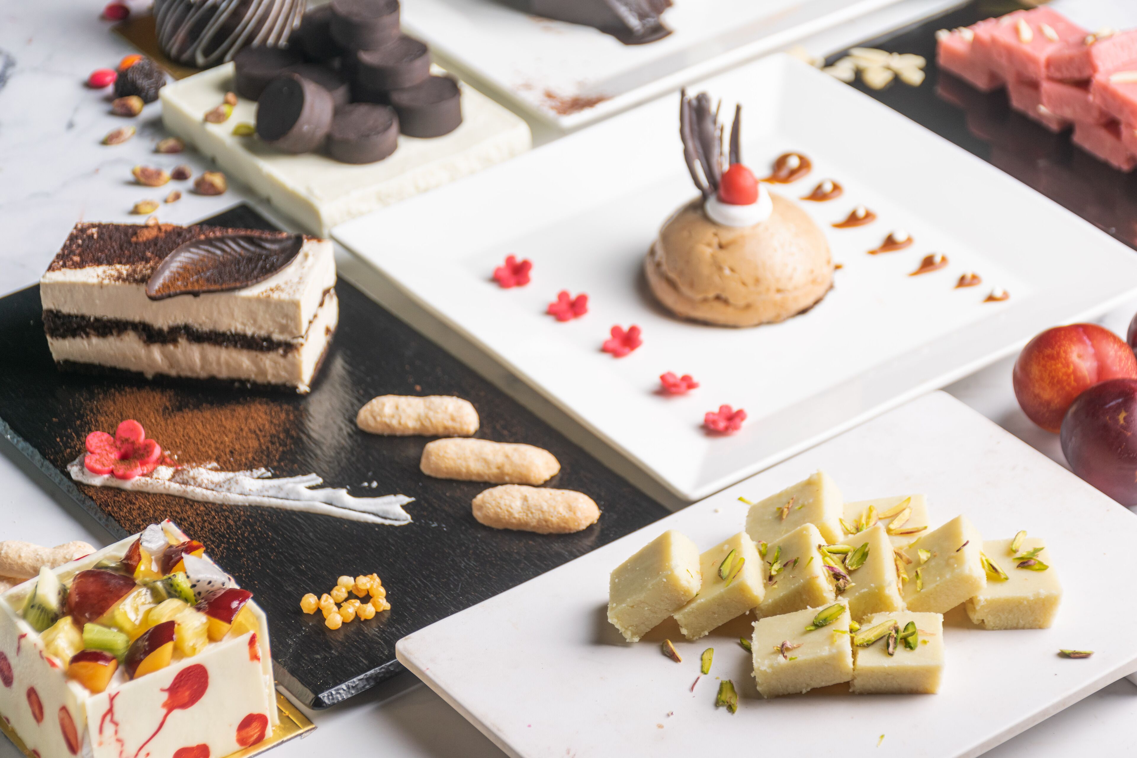 A variety of desserts on a table, including a layered cake, a round dessert with a cherry on top, and several square pieces of a white dessert garnished with nuts.