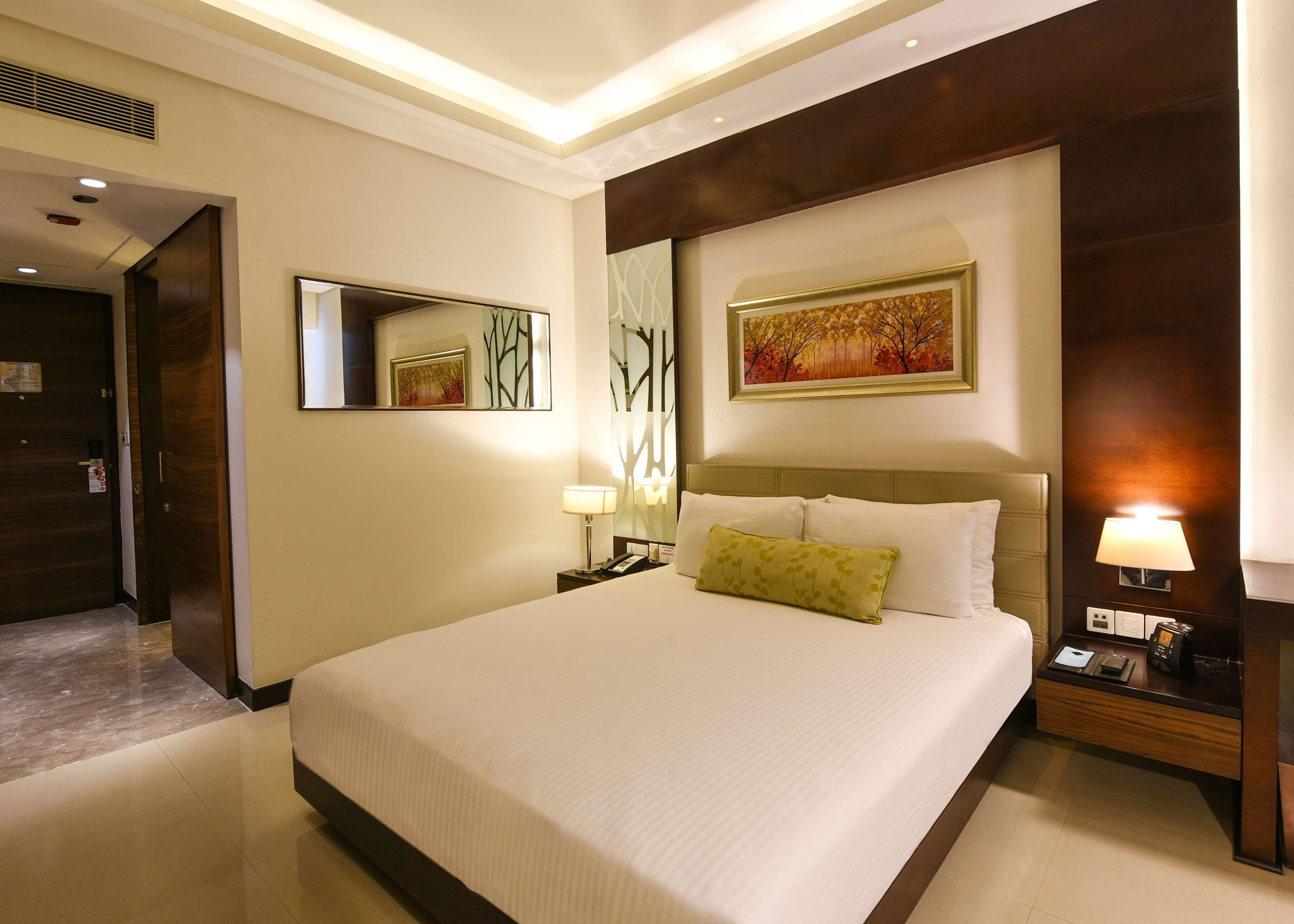 A hotel room with a large bed, a nightstand with a lamp, a bedside table with a clock, a mirror, and a painting on the wall.