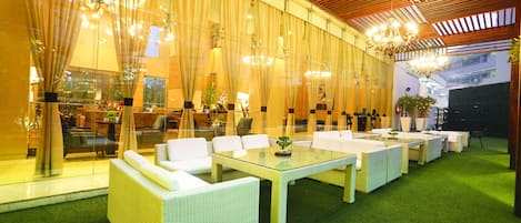 Un amplio espacio exterior con mobiliario de mimbre blanco y una mesa de vidrio, rodeado de cortinas translúcidas y una estructura de techo de madera.