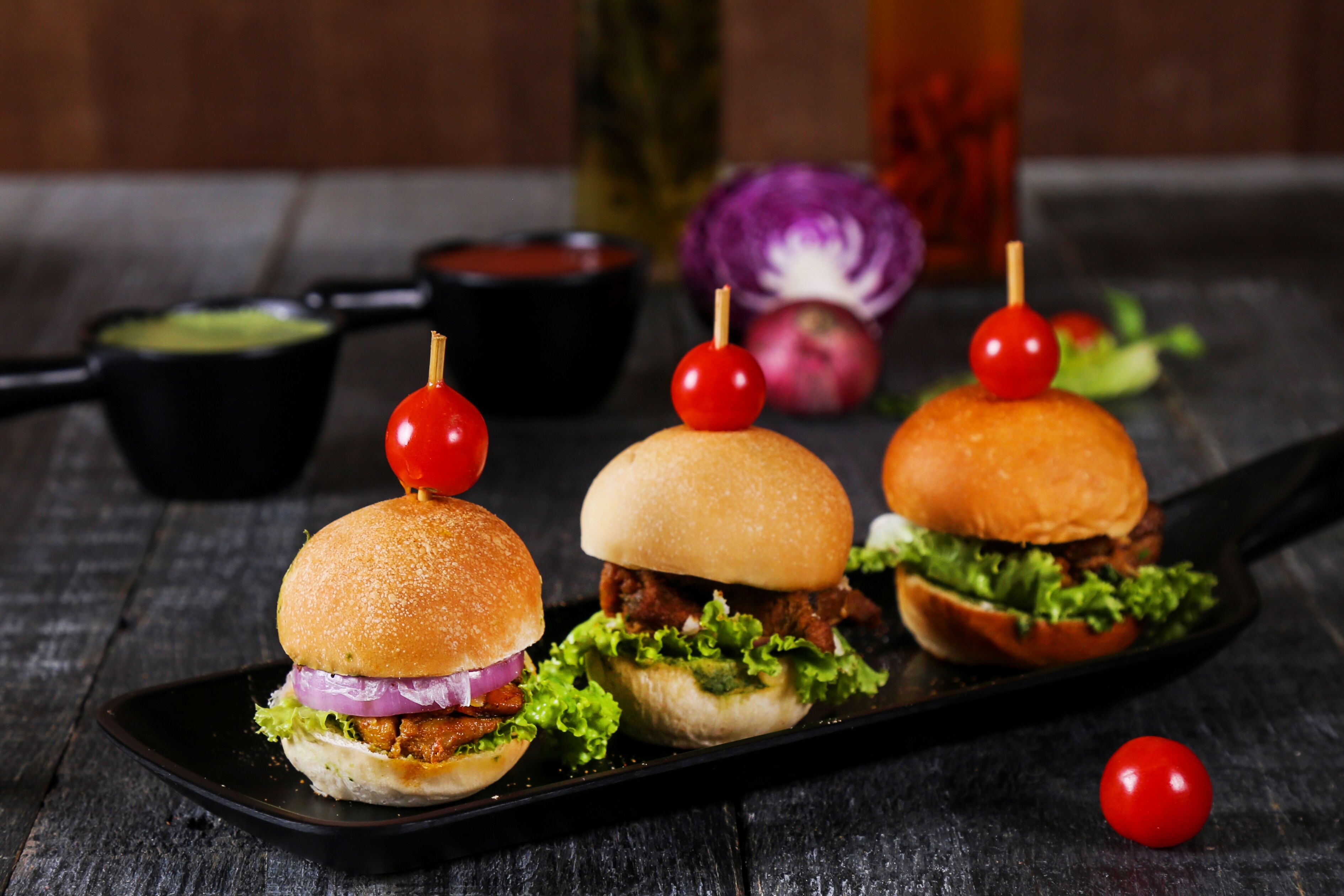 Trois hamburgers garnis de tomates cerises, servis sur une assiette noire avec une portion de sauce et des légumes en accompagnement.