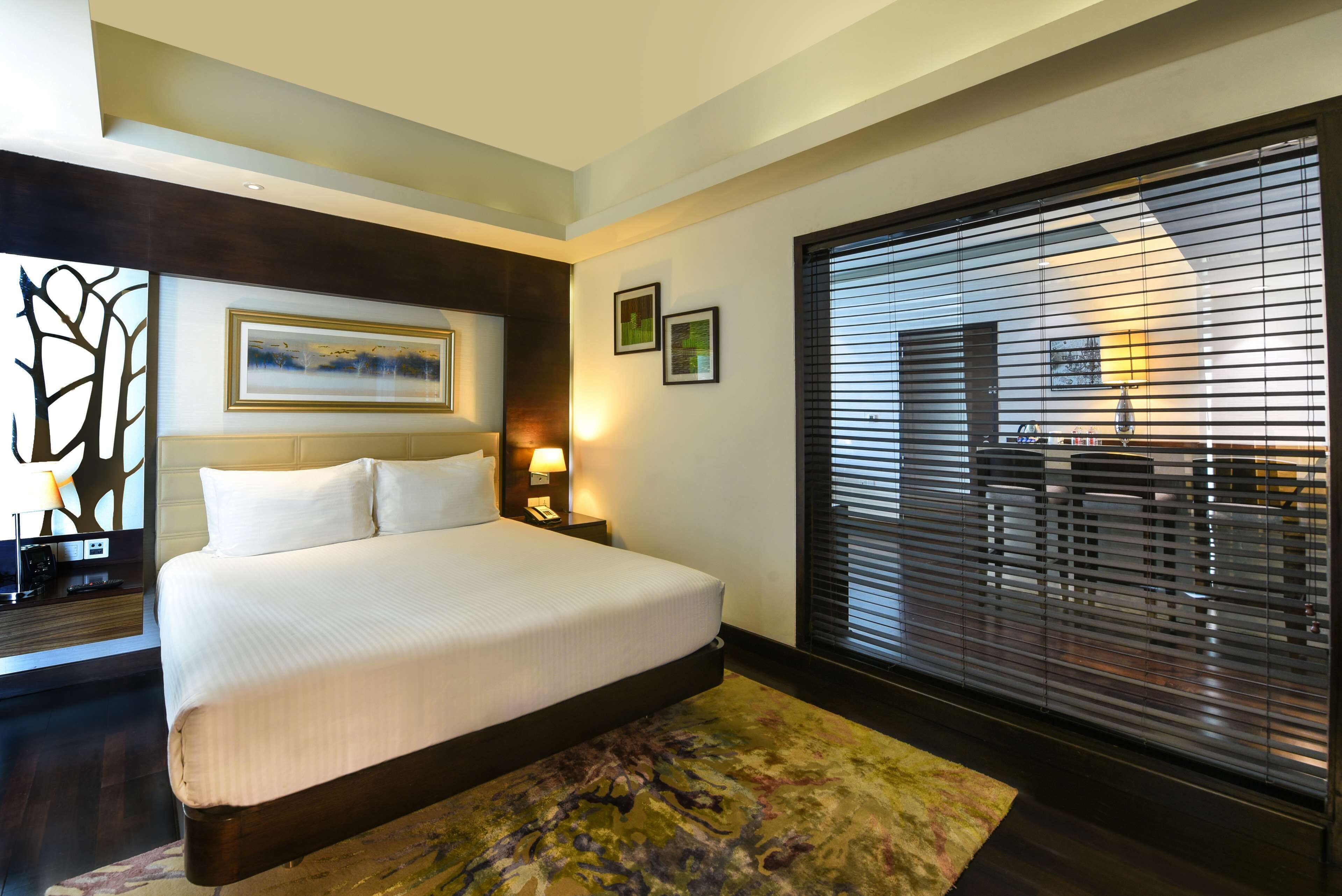 A hotel room with a large bed, a nightstand with a lamp, a painting on the wall, and a window with blinds.