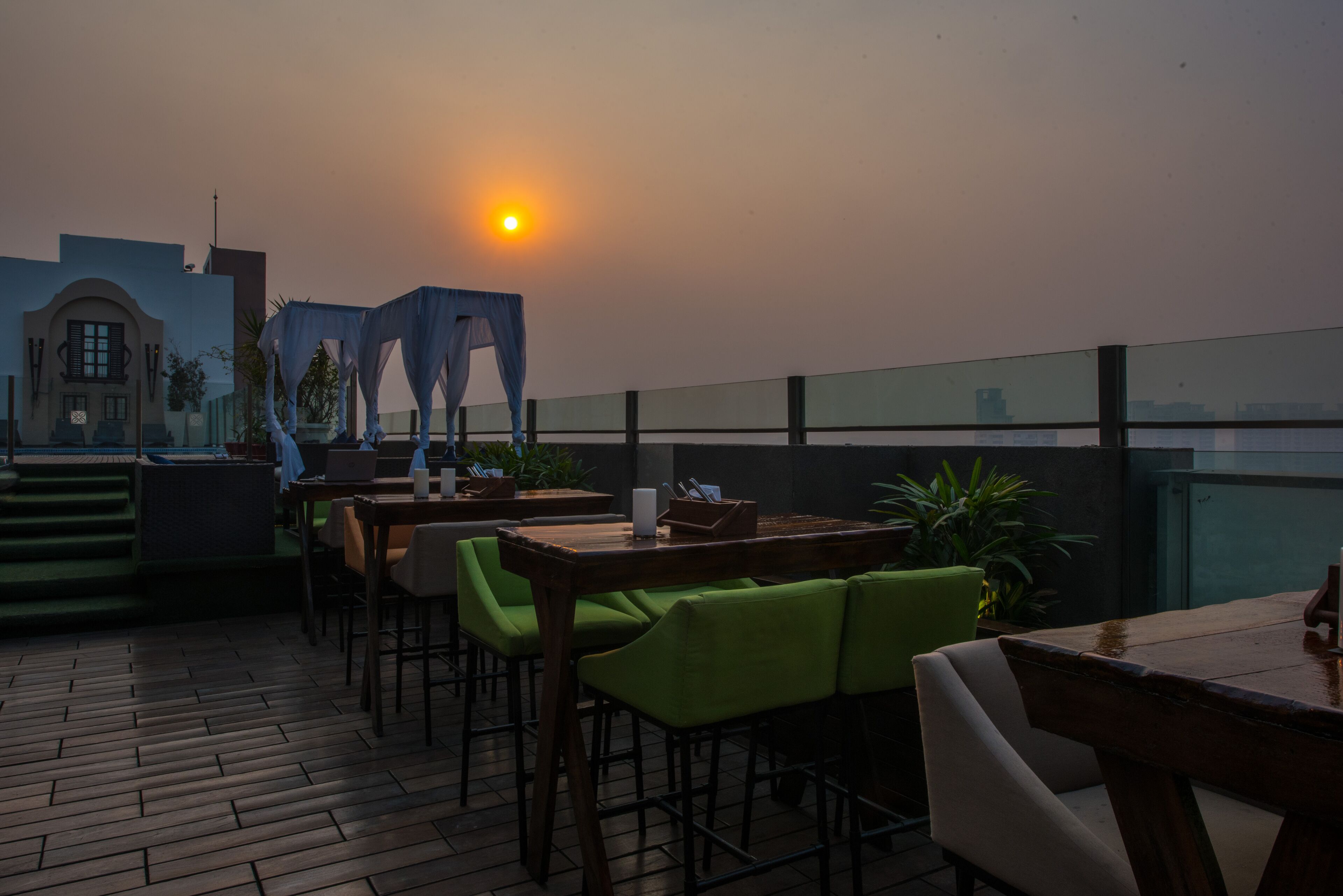 An outdoor seating area with wooden tables and chairs, a small stage with a canopy, and a view of the city skyline in the background.
