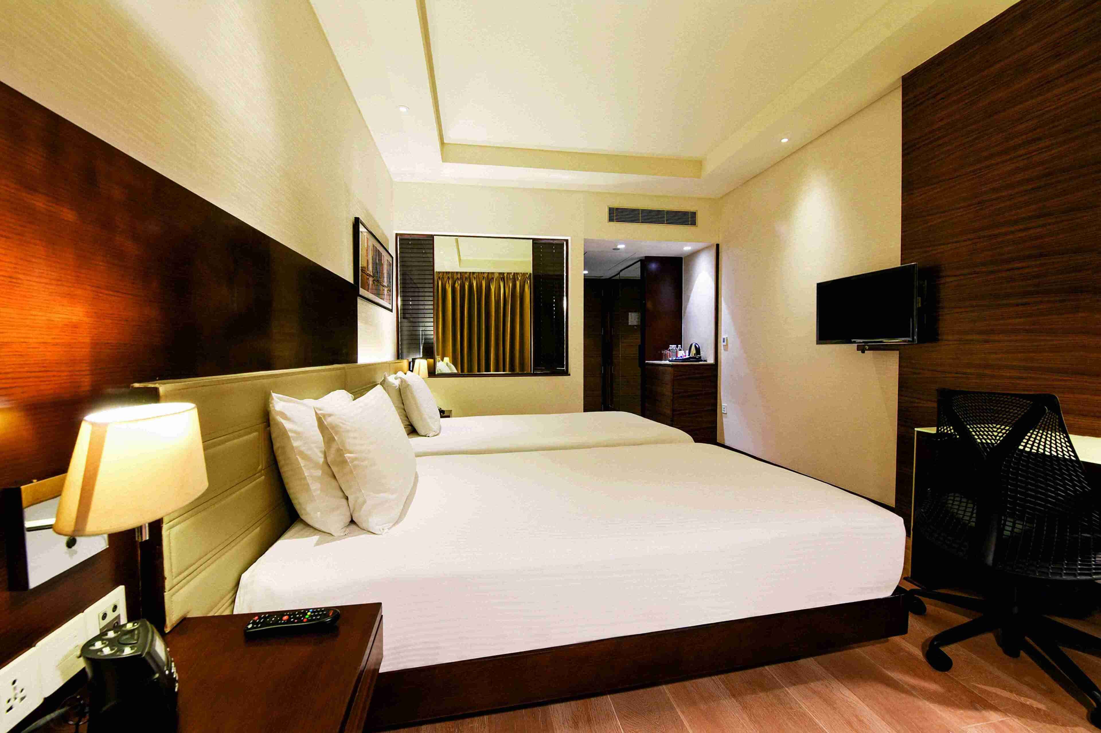 A hotel room with a large bed, a desk with a lamp, a chair, a TV mounted on the wall, and a bathroom visible through an open door.