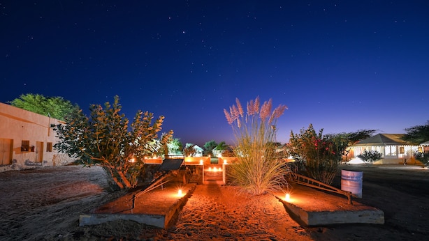 Vista nocturna de la entrada de un hotel, con un camino empedrado que lleva a la puerta, rodeada de zonas ajardinadas con plantas y un cielo despejado.