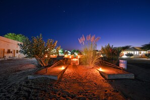 Vista nocturna de la entrada de un hotel, con un camino empedrado que lleva a la puerta, rodeada de zonas ajardinadas con plantas y un cielo despejado.
