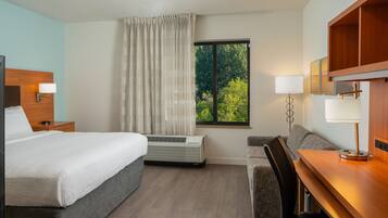 A hotel room with a bed, a desk, a chair, an air conditioning unit, and a window with a view of trees.