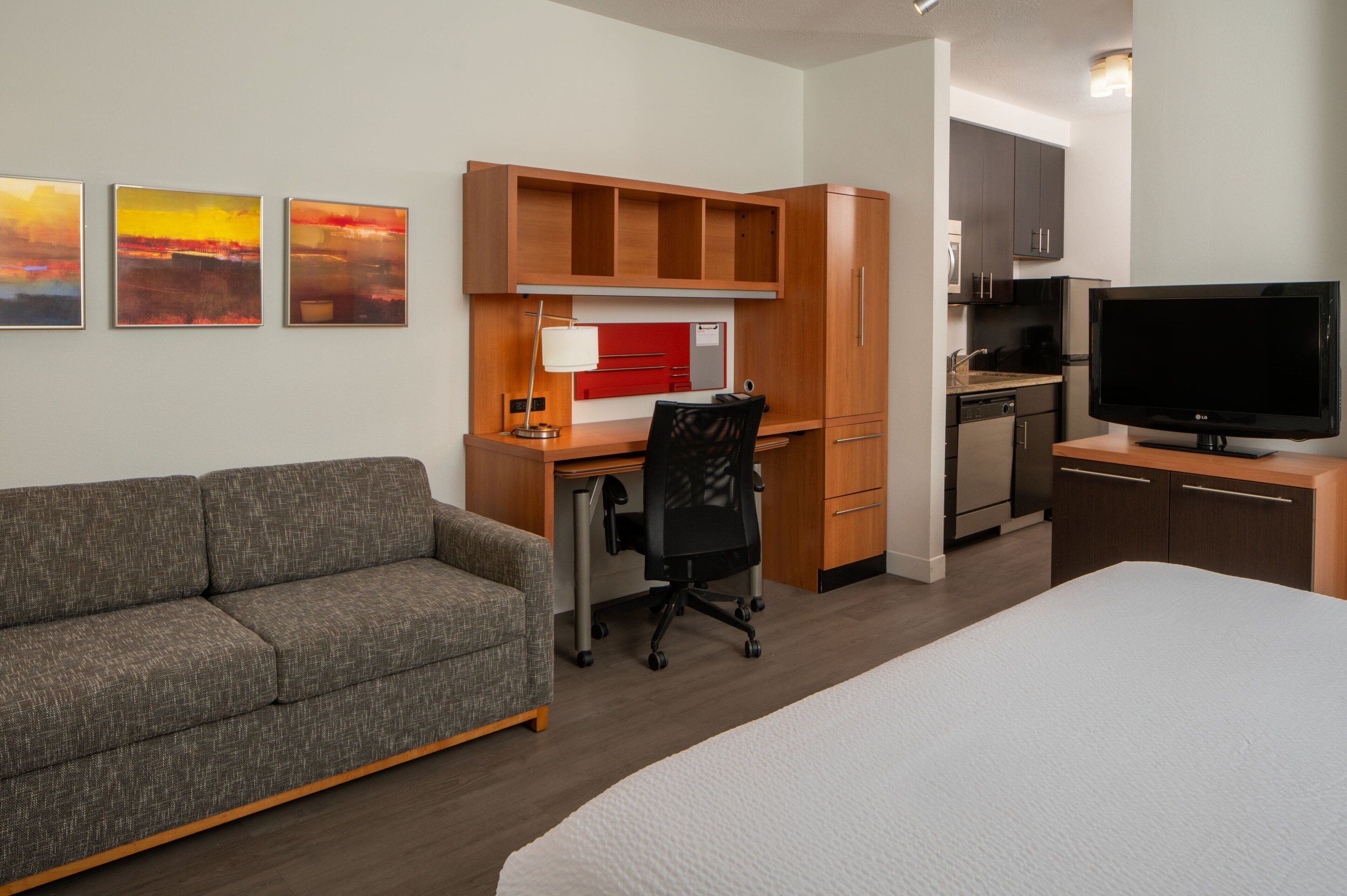 A hotel room with a bed, a sofa, a desk with a chair, a television, a kitchenette, and three paintings on the wall.