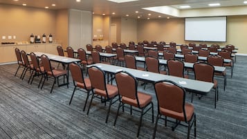 Uma sala de conferências com mesas e cadeiras dispostas em fileiras, uma tela de projetor e uma pequena copa.
