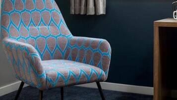 Une chaise ornée d’un motif géométrique, une fenêtre avec des rideaux et une petite table en bois sur laquelle se trouve une tasse.