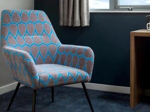 A chair with a geometric pattern, a window with curtains, and a small wooden table with a cup. - Travelodgellanelli Cross Hands (Llanelli)