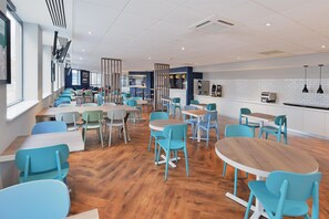 A modern dining area with wooden tables and teal chairs, a kitchen area with a white tiled backsplash, and a large window providing natural light. - Travelodge Woking Central (Woking)