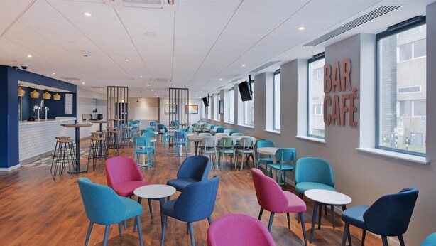 A modern bar cafe with a mix of blue and pink chairs, wooden tables, and a bar area with stools. - Travelodge Woking Central (Woking)