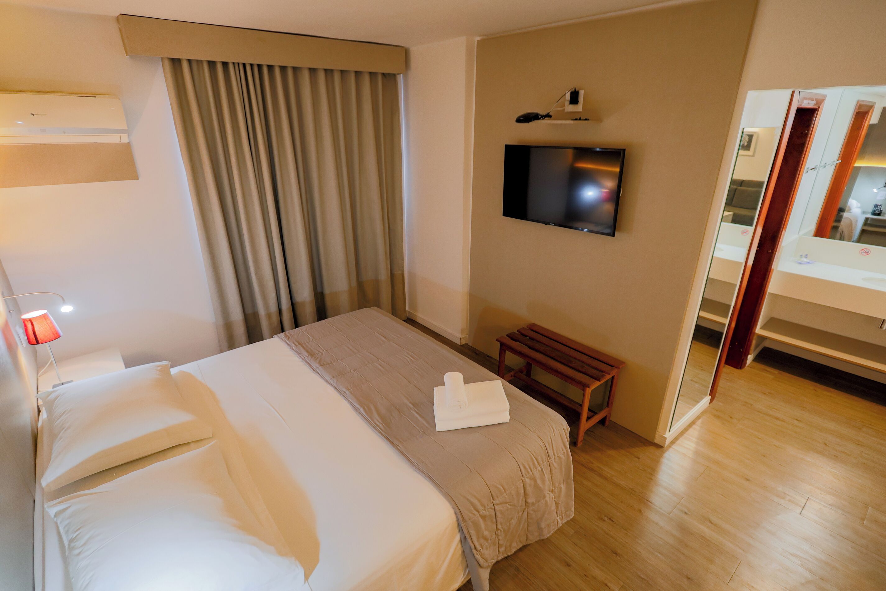Una habitación de hotel con una cama, un televisor montado en la pared, un banco de madera y un espejo.