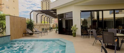 Un área de piscina moderna al aire libre con jacuzzi, sillones de descanso y un patio con mesas y sillas.