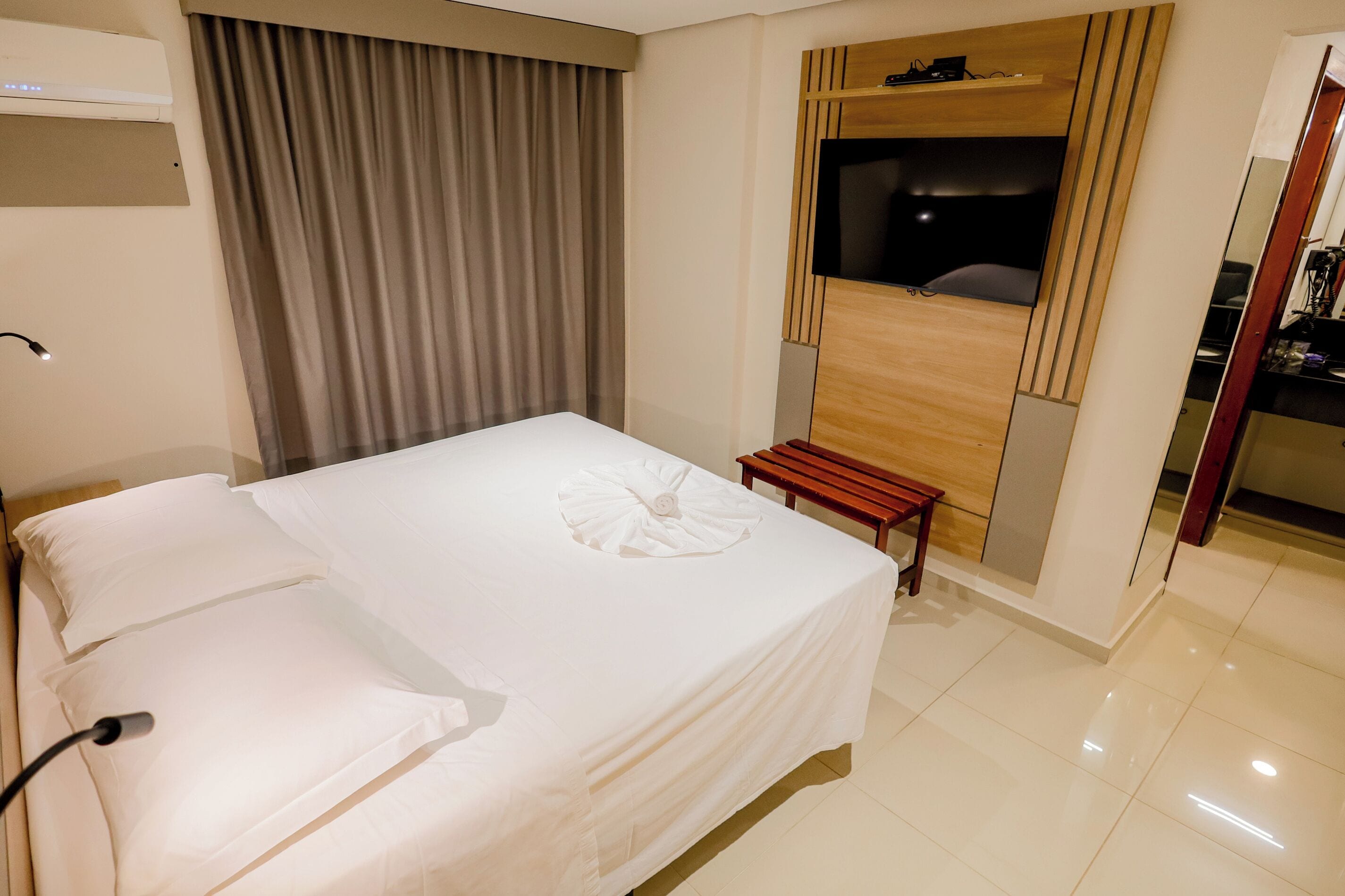 Ein Hotelzimmer mit einem großen Bett, einer hölzernen Sitzbank, einem an der Wand befestigten Fernseher und einem Spiegel.