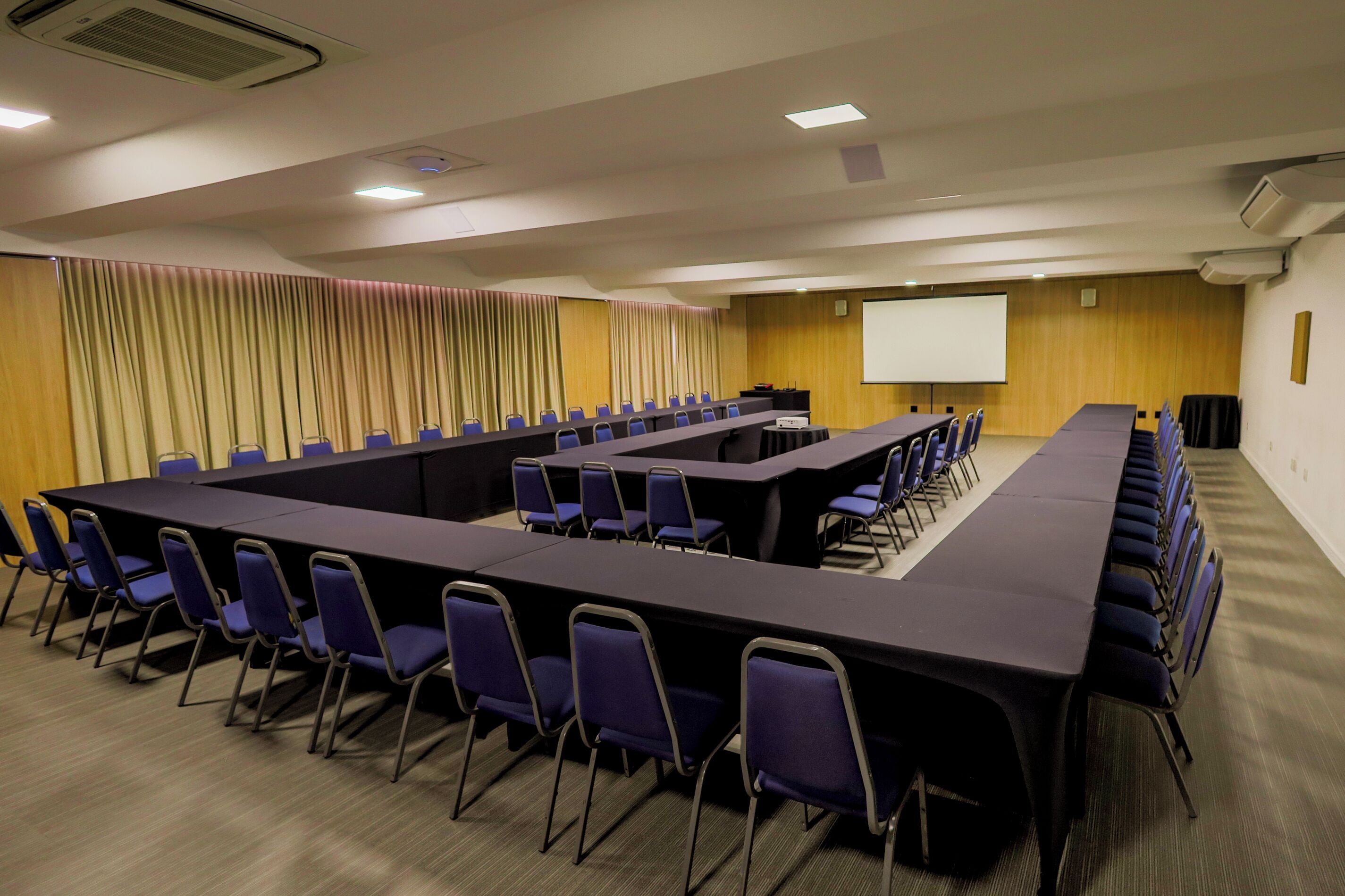 Un salón de conferencias con largas mesas y sillas, una pantalla de proyector y un revestimiento de pared de madera.