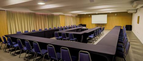 Un salón de conferencias con largas mesas y sillas, una pantalla de proyector y un revestimiento de pared de madera.