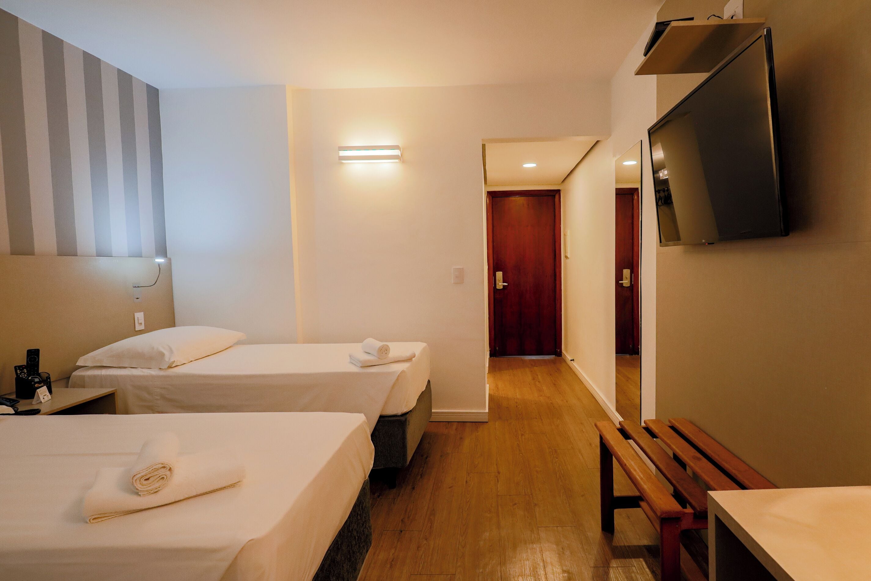 Habitación de hotel con dos camas, un banco de madera, un televisor montado en la pared y un pasillo que conduce a otra habitación.