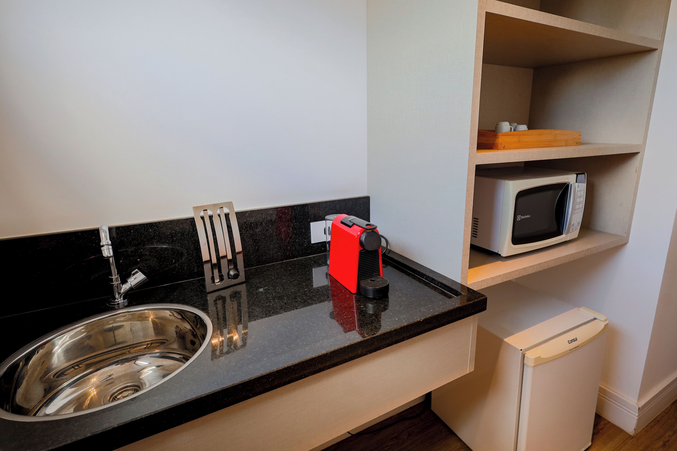 Una cocina compacta con un lavamanos de acero inoxidable, una cafetera roja, un microondas y un refrigerador pequeño.