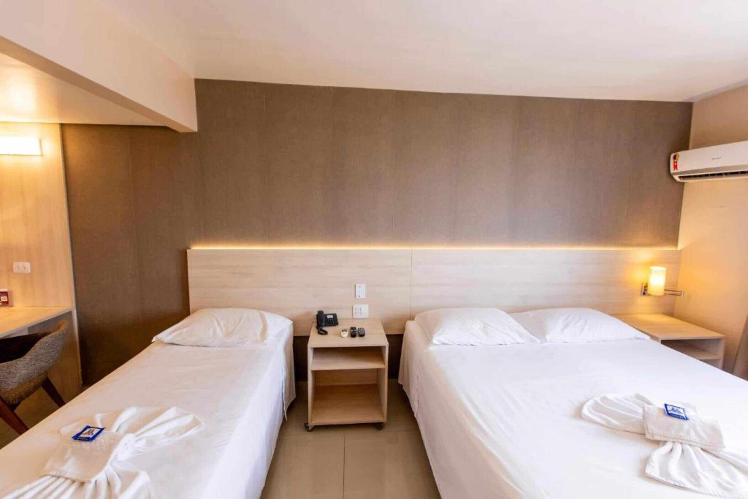 Quarto de hotel com duas camas de solteiro, cabeceira de madeira, mesas de cabeceira com luminárias e ar condicionado fixado na parede.