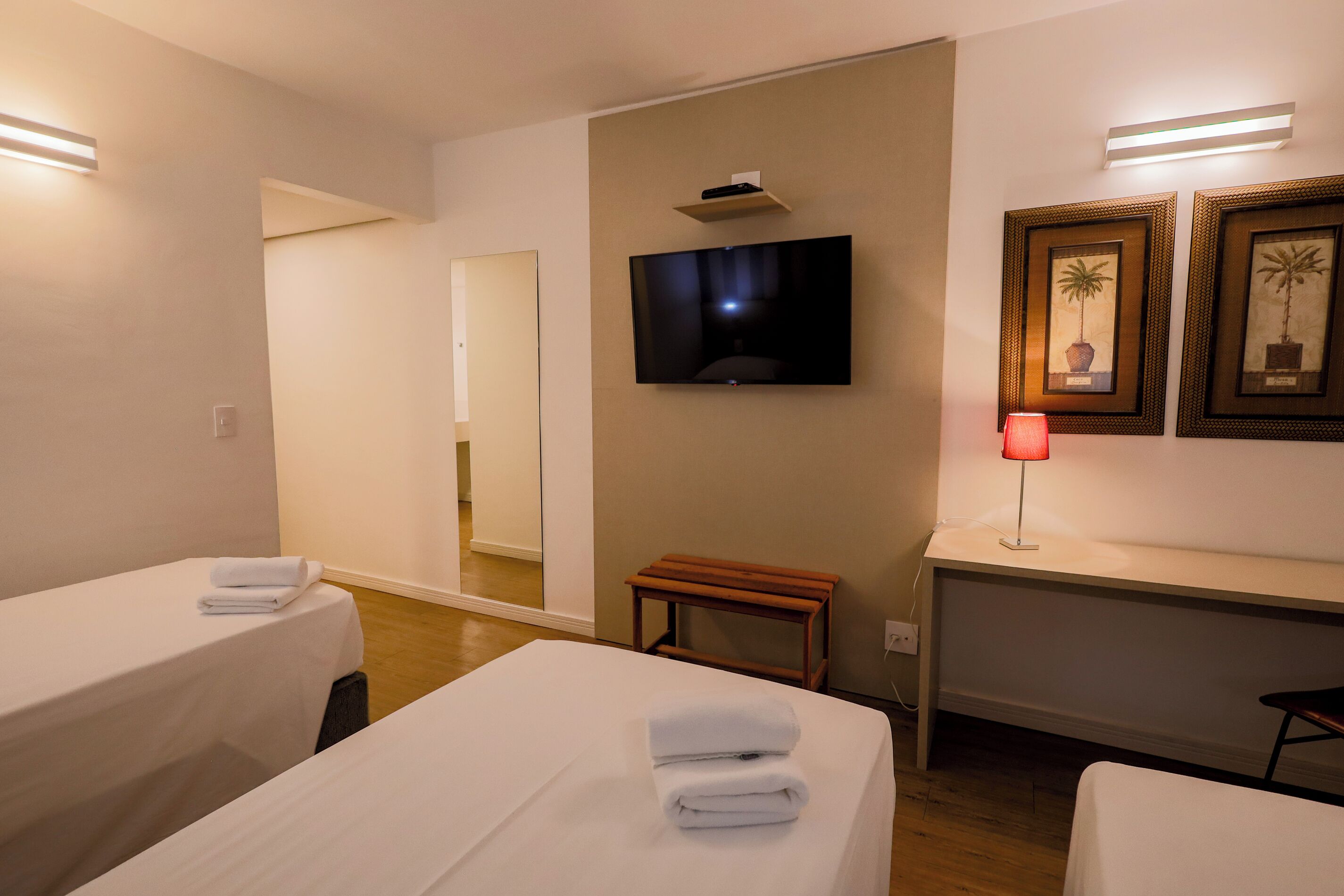 Habitación de hotel con dos camas, un televisor montado en la pared, una mesita y cuadros enmarcados.