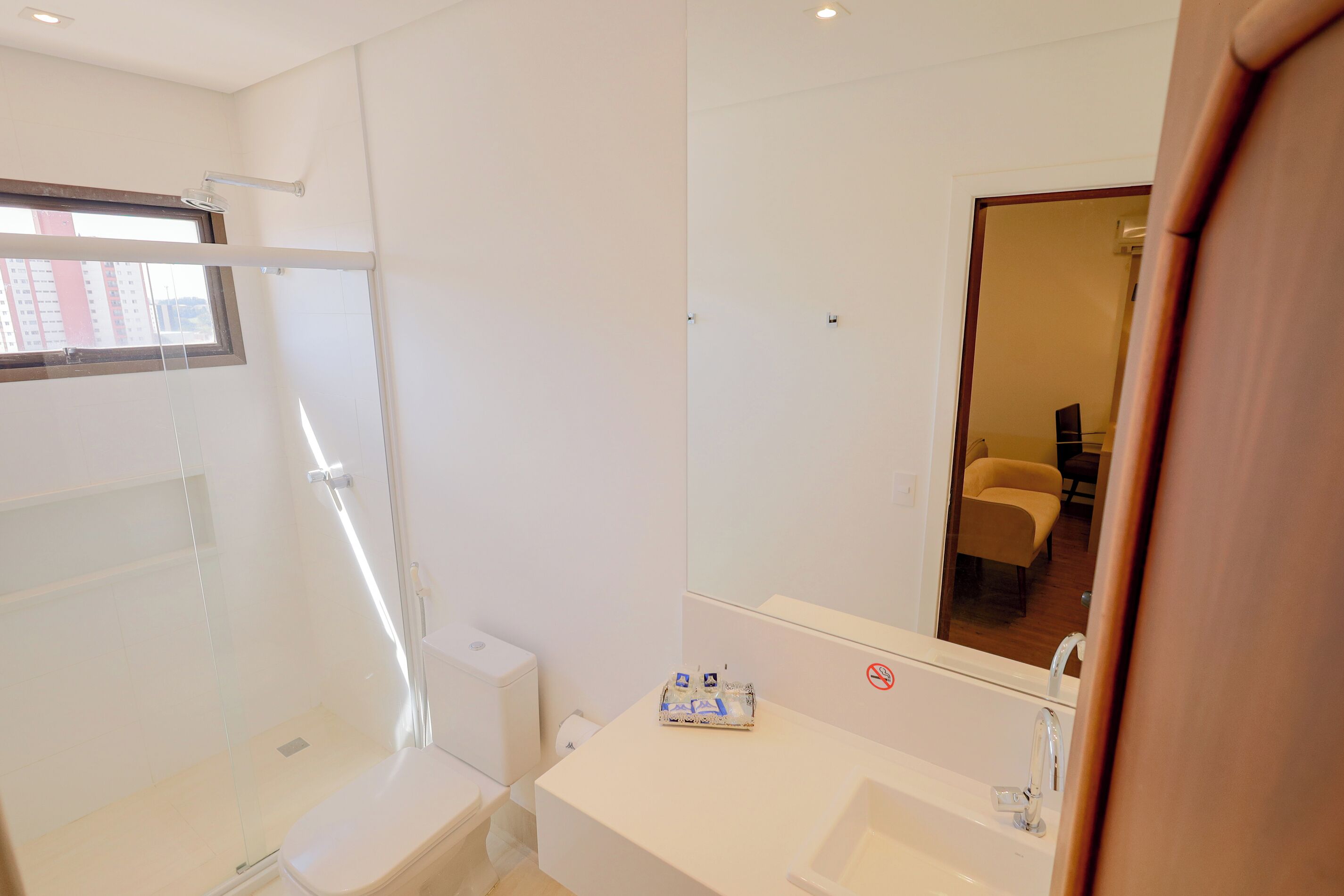 Une salle de bain moderne avec une douche à parois de verre, un lavabo blanc et un miroir qui reflète une chaise jaune.