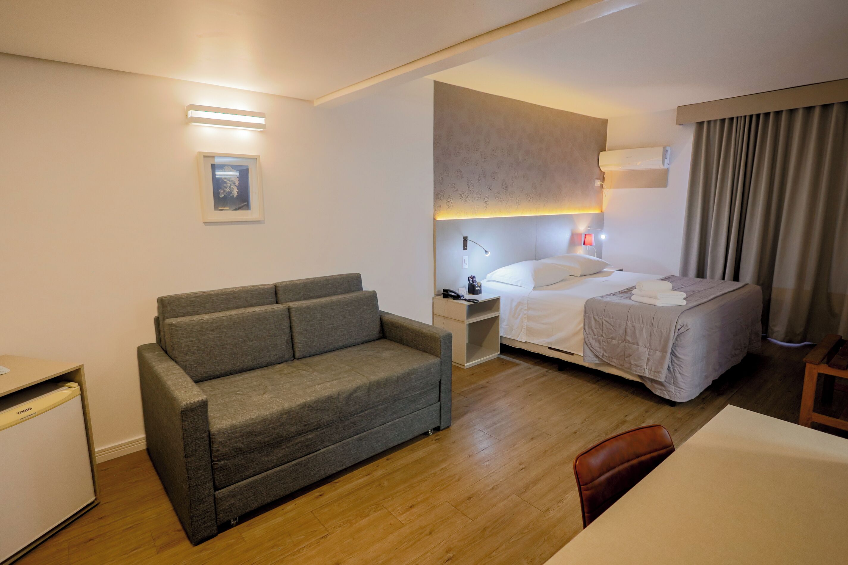 Habitación de hotel con sofá gris, cama con edredón blanco, mesita de noche con lámpara y un escritorio pequeño con una computadora.