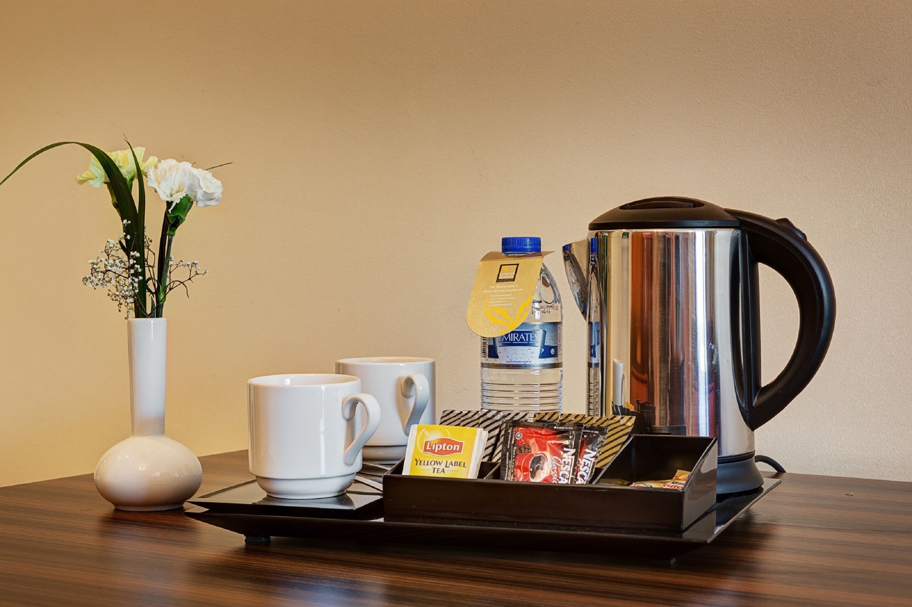 Un desayuno con una cafetera, dos tazas blancas, un florero con flores, una botella de agua y una bandeja con sales de té.