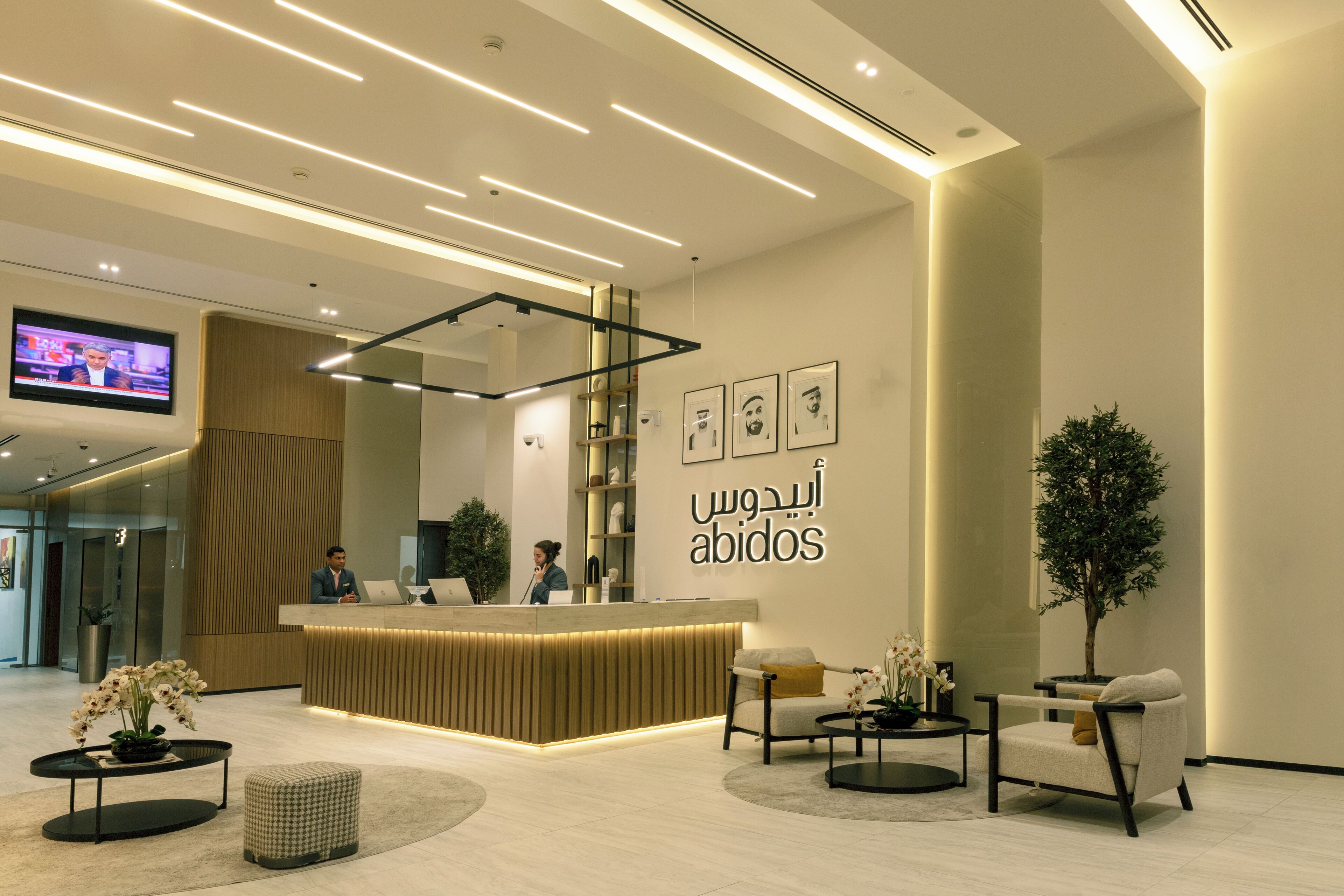A modern hotel reception area with a wooden reception desk, a flat-screen TV, and a wall with framed portraits.