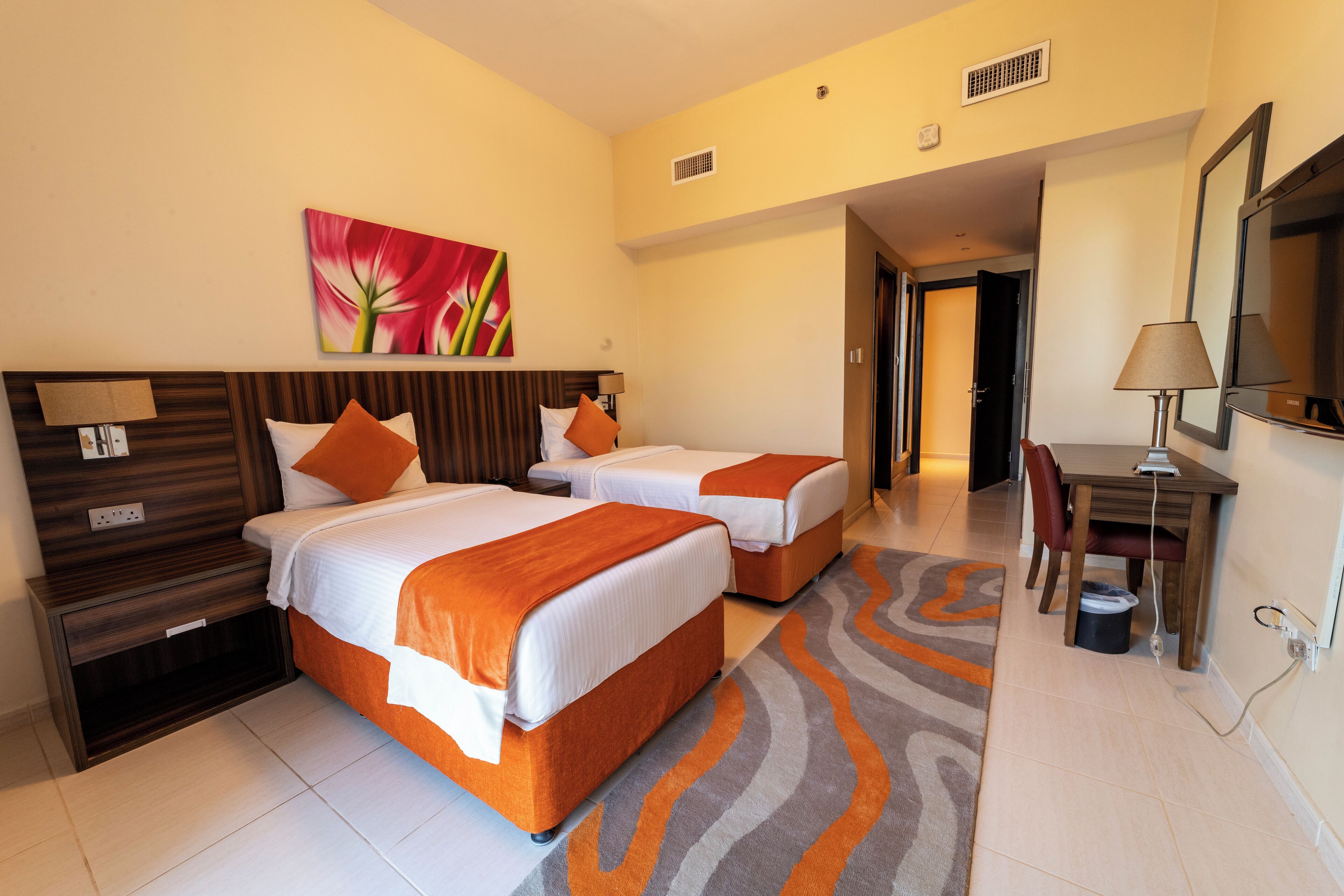 A hotel room with two beds, a desk, a chair, a painting, and a bathroom door.