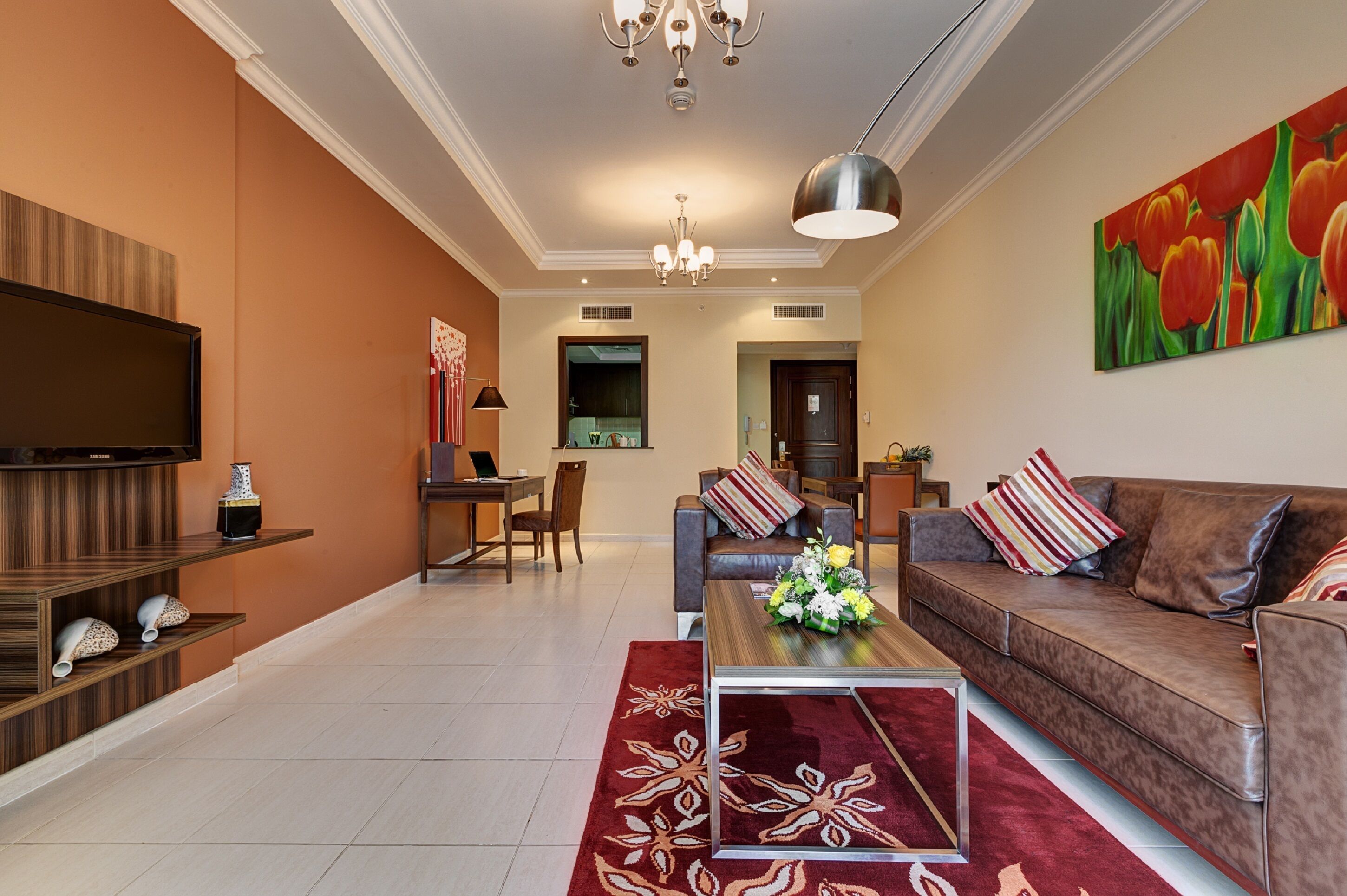 A modern living room with a flat-screen TV, a sofa set, a coffee table with flowers, and a red patterned rug.