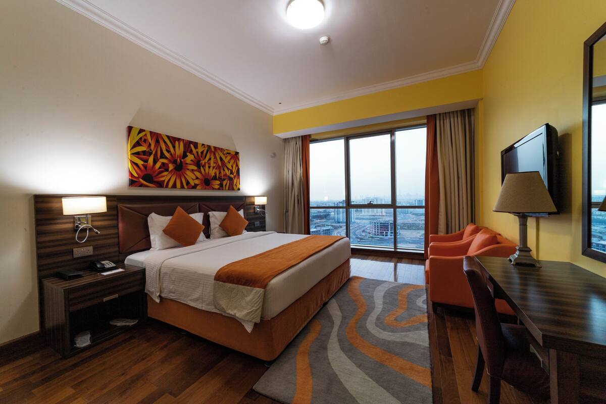 Habitación de hotel con una cama grande, un escritorio, una silla, un televisor y vista a la ciudad.